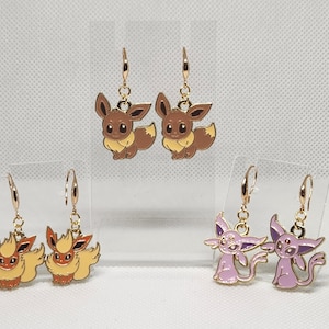 Könnte beinhalten: Goldfarbene Ohrhänger mit Cartoon-Charakteren aus braunem, orangefarbenem und lila Emaille. Die Ohrringe zeigen drei verschiedene Charakterdesigns, jedes mit einer einzigartigen Farbpalette und Stil, ideal für Fans der Franchise.