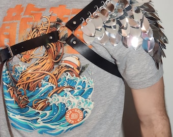 May include: Silver metal shoulder armour with a black leather harness. The armour is made of overlapping, heart-shaped scales. The person is wearing a grey t-shirt with a graphic of a dragon and waves.