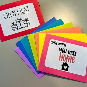 May include: A set of colorful envelopes with the text "Open First" on the top envelope and "Open When... You Miss Home" on the bottom envelope. The envelopes are arranged in a rainbow pattern.