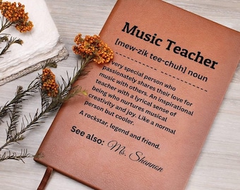 May include: A brown leather-bound notebook with the words "Music Teacher" and a definition printed on the cover. The definition describes a music teacher as an inspirational person. The notebook has a brown ribbon bookmark.