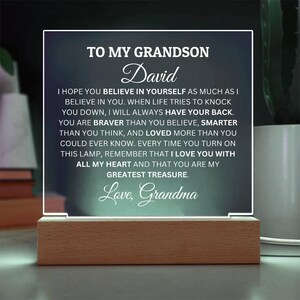 May include: A white acrylic plaque with a wooden base, featuring a message for a grandson named David. The message reads, "To my grandson David, I hope you believe in yourself as much as I believe in you. When life tries to knock you down, I will always have your back. You are braver than you believe, smarter than you think, and loved more than you could ever know. Every time you turn on this lamp, remember that I love you with all my heart and that you are my greatest treasure. Love, Grandma."