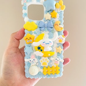 May include: A blue and white phone case with a whimsical design featuring a sleeping dog, clouds, stars, and a yellow fence. The case is decorated with 3D elements and has a cutout for the phone's camera.