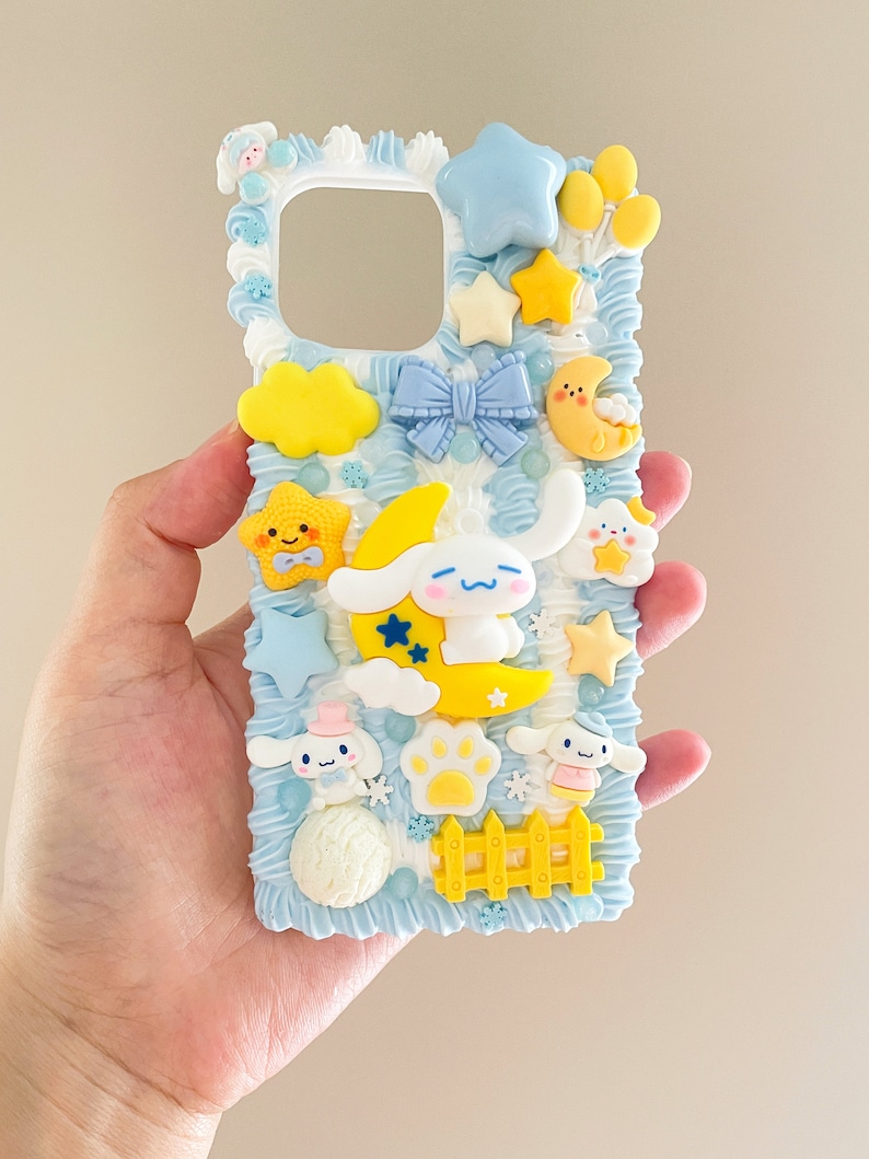 May include: A blue and white phone case with a whimsical design featuring a sleeping dog, clouds, stars, and a yellow fence. The case is decorated with 3D elements and has a cutout for the phone's camera.
