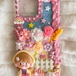 Pu&ograve; includere: Una custodia per telefono rosa e blu decorata con fiori 3D, una staccionata bianca, una bambina dei cartoni animati e un cartello che dice "Credi nei sogni".