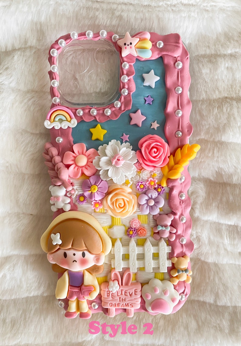 Pu&ograve; includere: Una custodia per telefono rosa e blu decorata con fiori 3D, una staccionata bianca, una bambina dei cartoni animati e un cartello che dice "Credi nei sogni".