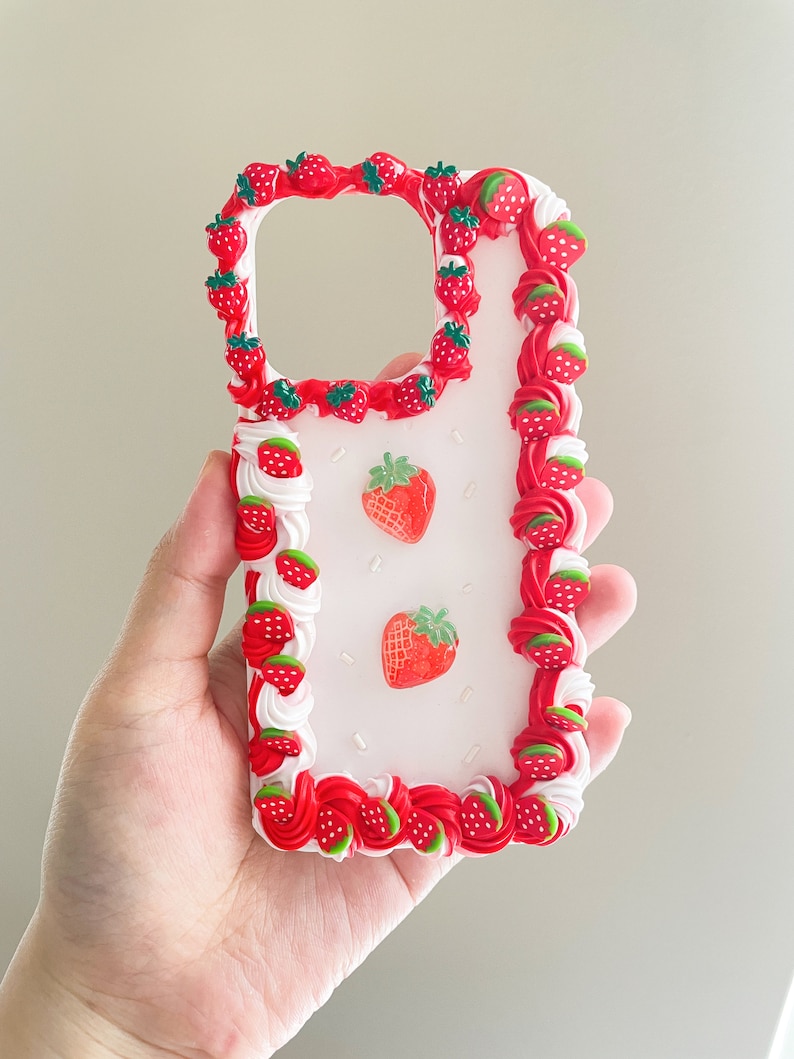 May include: A white phone case with a red and white frosting design and strawberry decorations.