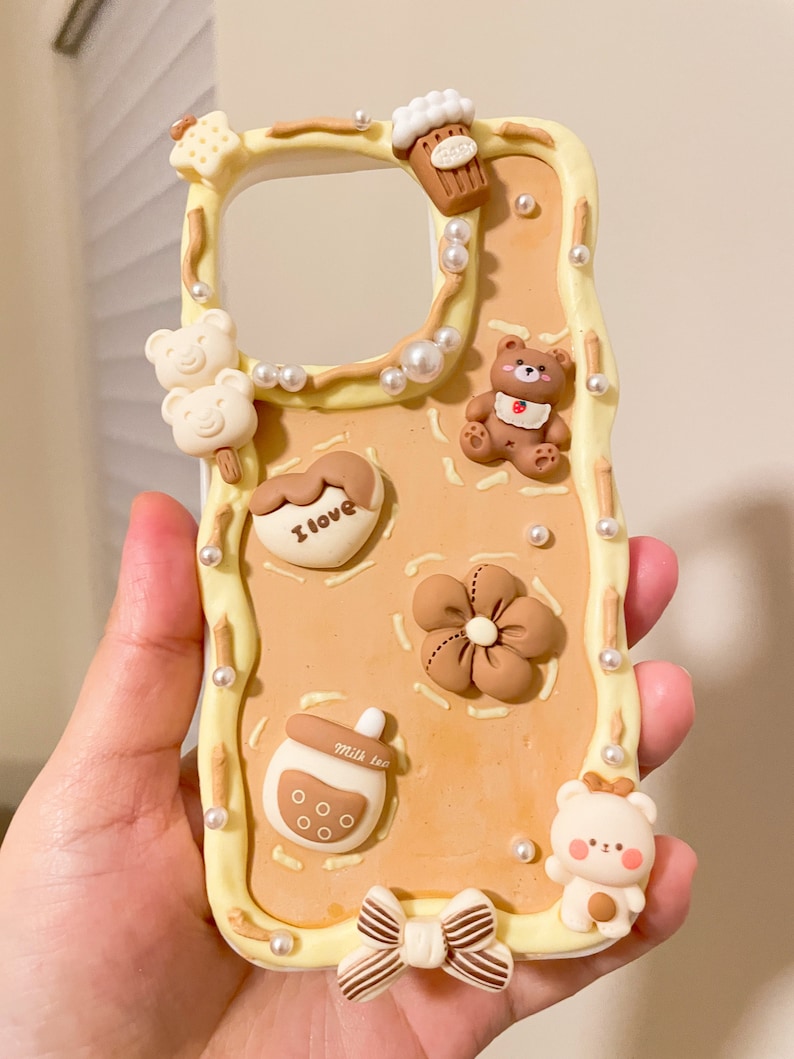 May include: A light brown phone case with a white border and various brown and white clay decorations. The decorations include a bear, a flower, a heart with the text "I love", a milk tea cup, a bow, and a star.