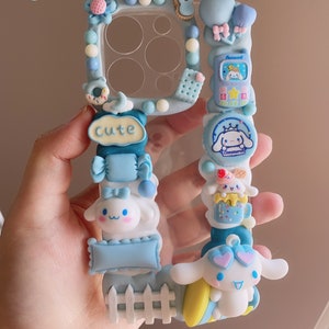 May include: A blue and white phone case decorated with a variety of 3D charms, including a small white dog with a pink bow, a blue and white candy cane, a blue and white fence, and a blue and white sign that says "Cute".