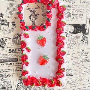 May include: A white phone case decorated with red and white frosting, strawberries, and green leaves. The case has a clear window in the center.