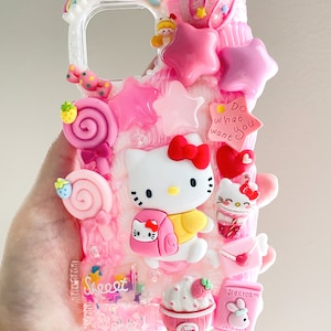 May include: A pink and white Hello Kitty phone case with a glittery background. The case features a 3D Hello Kitty figure, a pink lollipop, a pink star, a pink heart, and a pink cupcake. The case also has the text "Do what you want" on it.