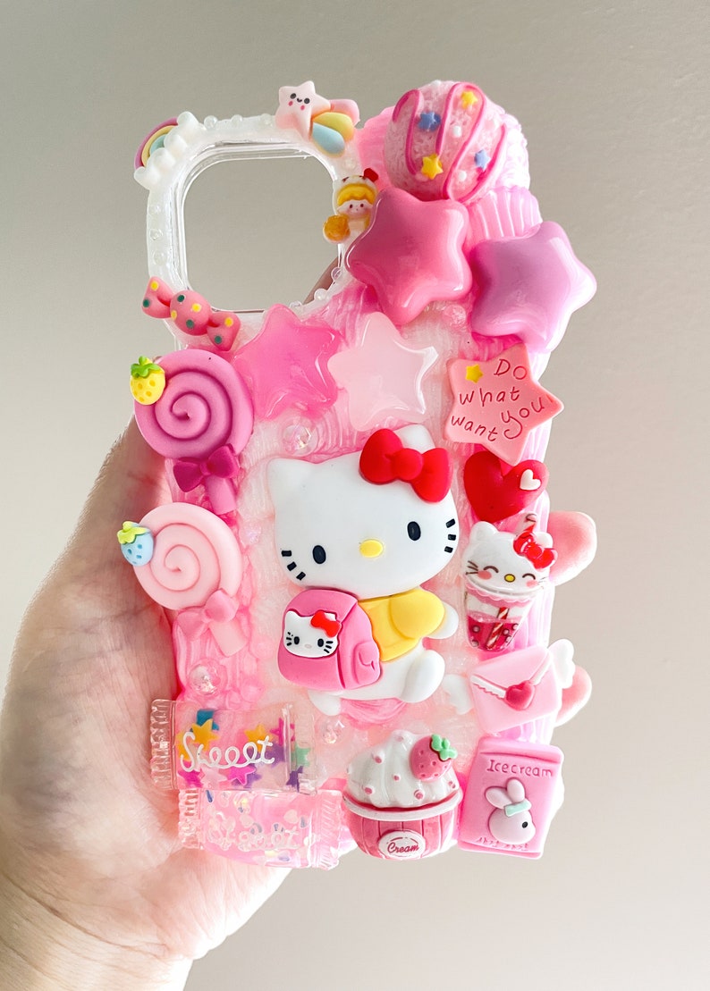 May include: A pink and white Hello Kitty phone case with a glittery background. The case features a 3D Hello Kitty figure, a pink lollipop, a pink star, a pink heart, and a pink cupcake. The case also has the text "Do what you want" on it.