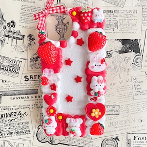 May include: A white phone case decorated with red, white, and pink 3D embellishments. The case features strawberries, hearts, stars, and bunnies. The case is surrounded by vintage advertisements.