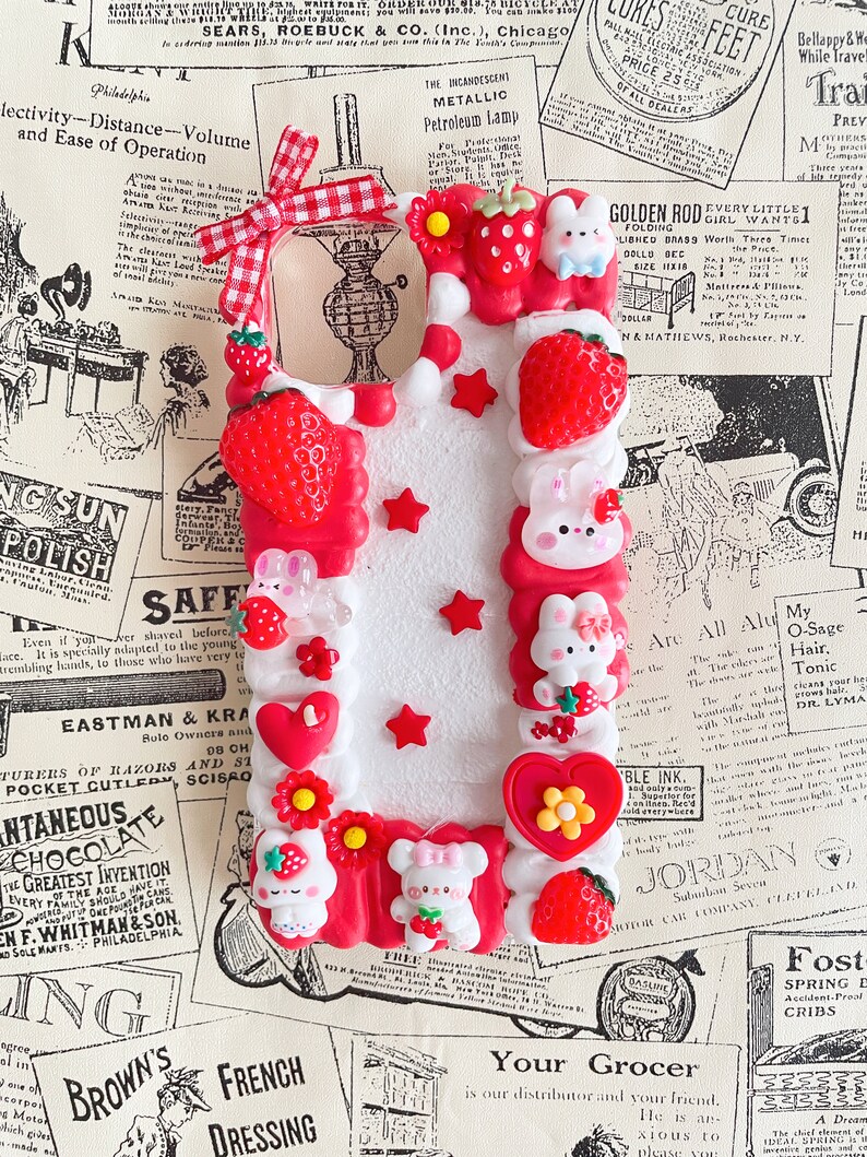 May include: A white phone case decorated with red, white, and pink 3D embellishments. The case features strawberries, hearts, stars, and bunnies. The case is surrounded by vintage advertisements.