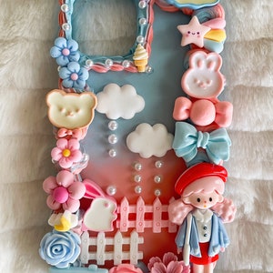 Pu&ograve; includere: Una custodia per telefono blu pastello e rosa decorata con decorazioni 3D, tra cui una bambola, un coniglio, nuvole, fiori, una recinzione, una stella, un arcobaleno e un cartello che dice "Rabbitz House". La custodia ha anche la scritta "Cute".