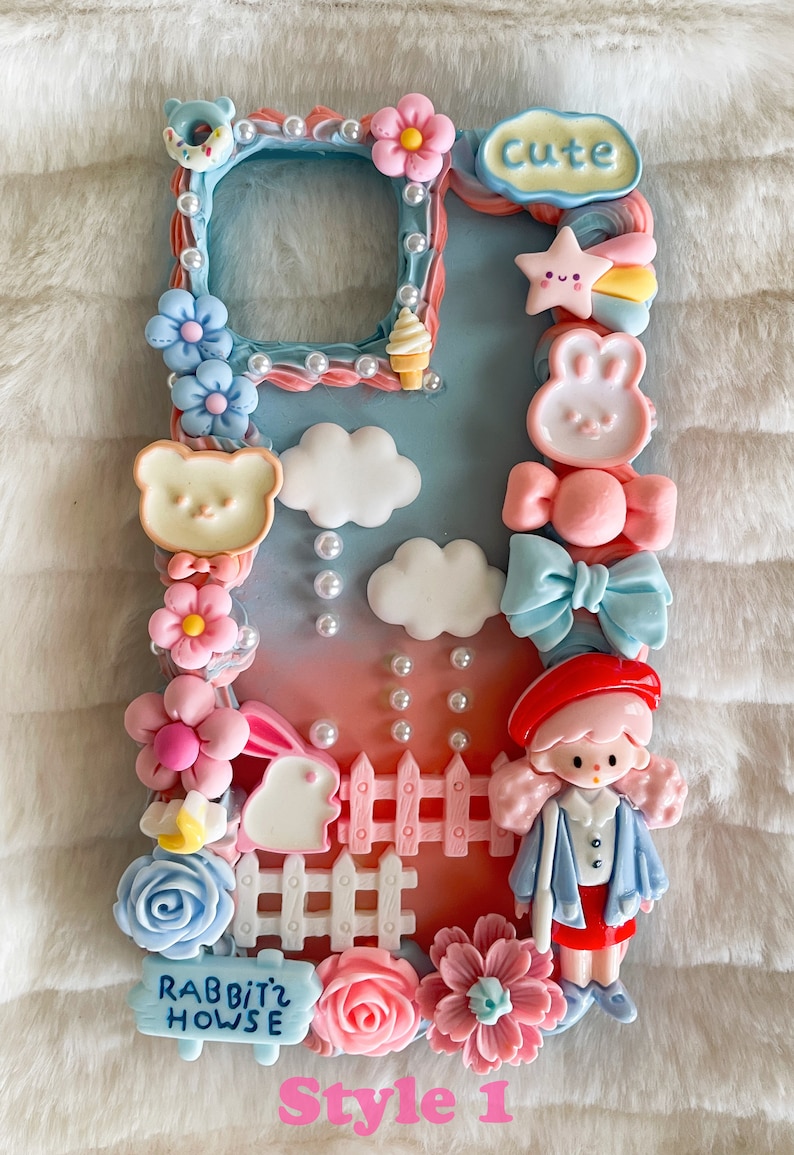 Pu&ograve; includere: Una custodia per telefono blu pastello e rosa decorata con decorazioni 3D, tra cui una bambola, un coniglio, nuvole, fiori, una recinzione, una stella, un arcobaleno e un cartello che dice "Rabbitz House". La custodia ha anche la scritta "Cute".