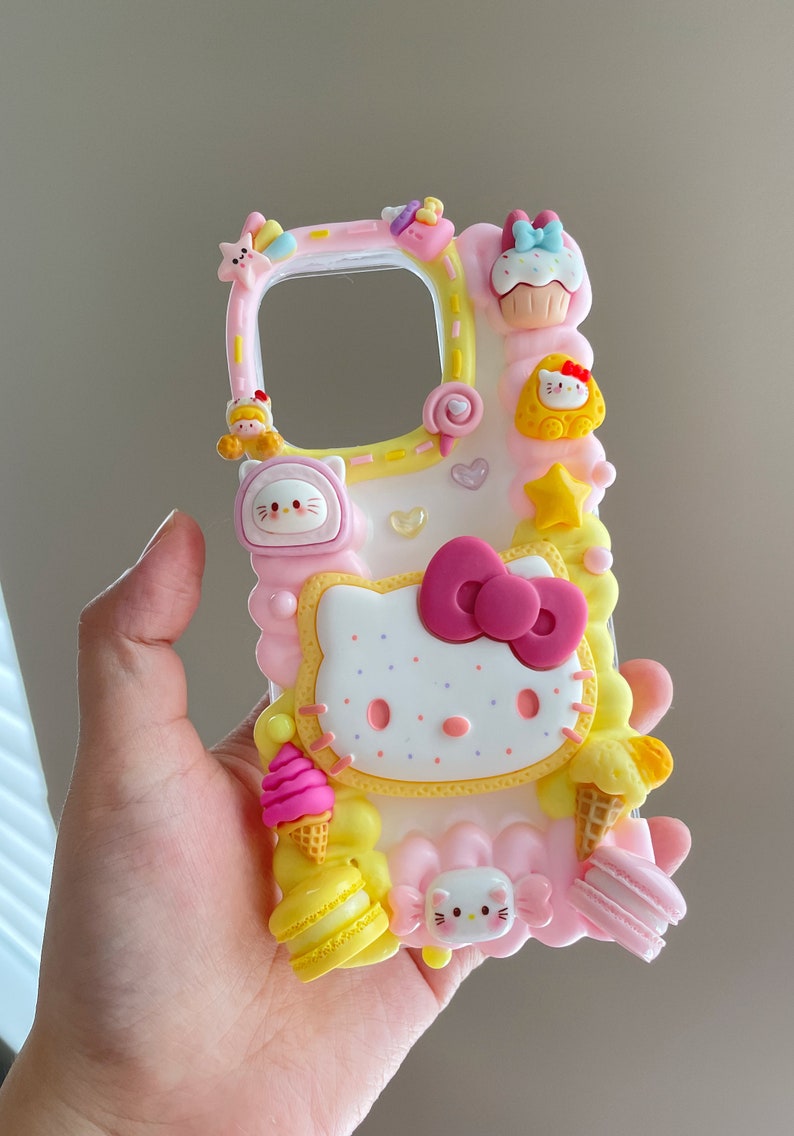 May include: A pink and yellow phone case featuring a Hello Kitty design. The case includes a Hello Kitty cookie, ice cream cones, macarons, and other cute details.