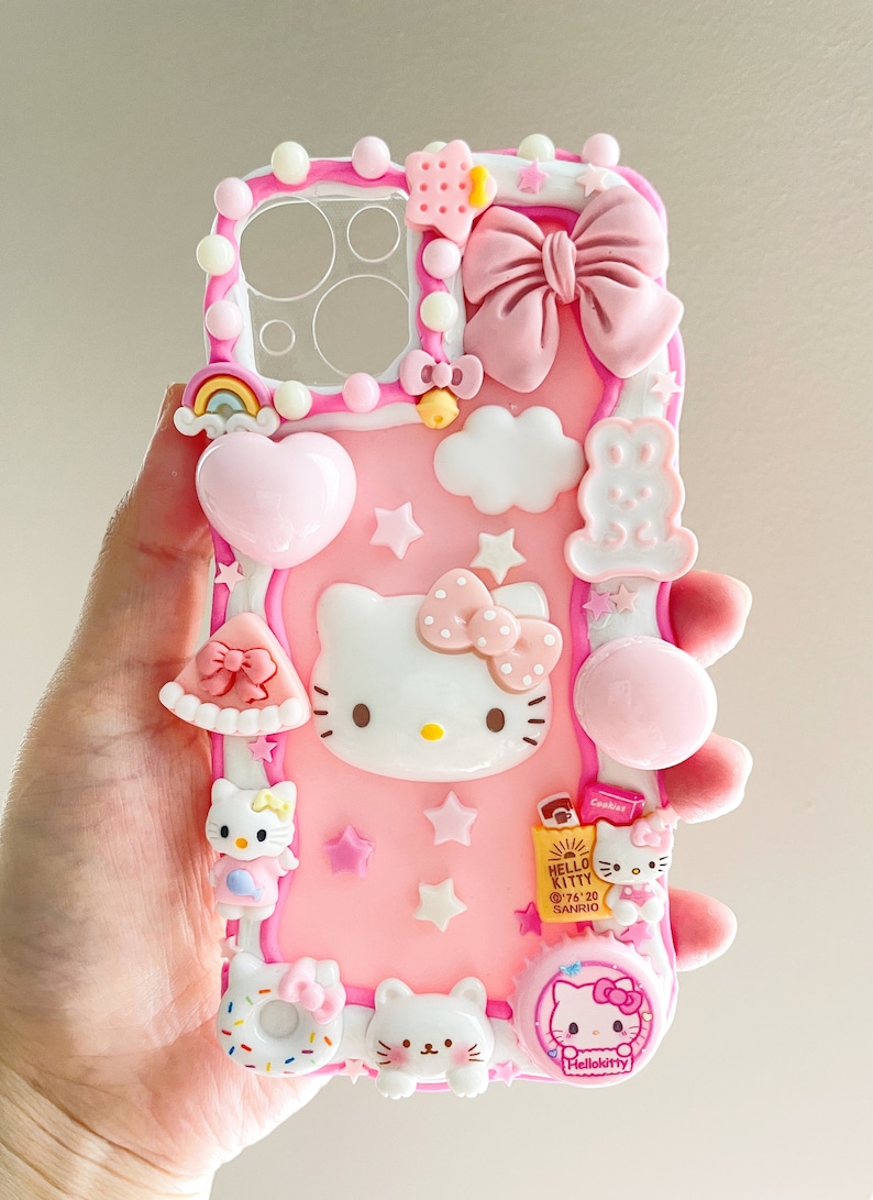 May include: A pink Hello Kitty phone case with 3D embellishments, including a bow, a cloud, a heart, stars, and Hello Kitty characters. The case has a clear back panel for the camera lens.