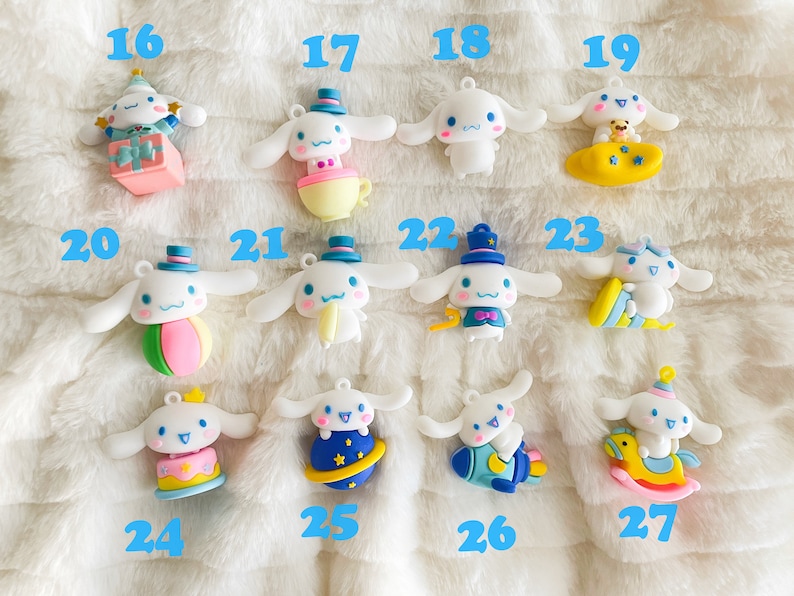 May include: A collection of 12 white plush dog charms with different colored accessories. The charms are numbered 16 through 27. Each charm features a different design, including a birthday cake, a ball, a teacup, a rocket, a rocking horse, and a planet.
