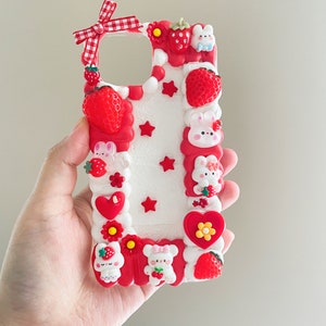 May include: A white phone case decorated with red and white strawberries, white bunnies, red hearts, and white stars. The case has a red bow with white checkered pattern.