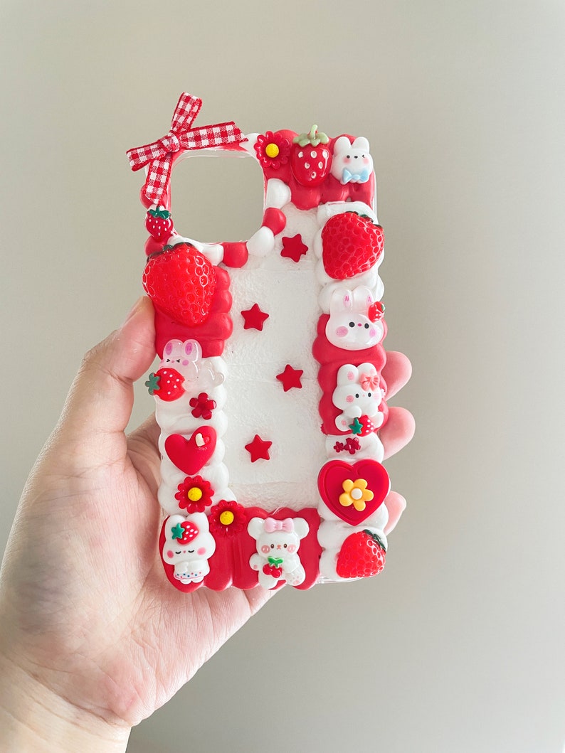 May include: A white phone case decorated with red and white strawberries, white bunnies, red hearts, and white stars. The case has a red bow with white checkered pattern.