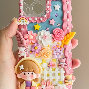 Pu&ograve; includere: Una custodia per telefono rosa e blu decorata con fiori, stelle, un arcobaleno, una recinzione, una ragazza, un coniglio e un'impronta di zampa. La custodia ha il testo "Believe in Dreams" su un cartello giallo.