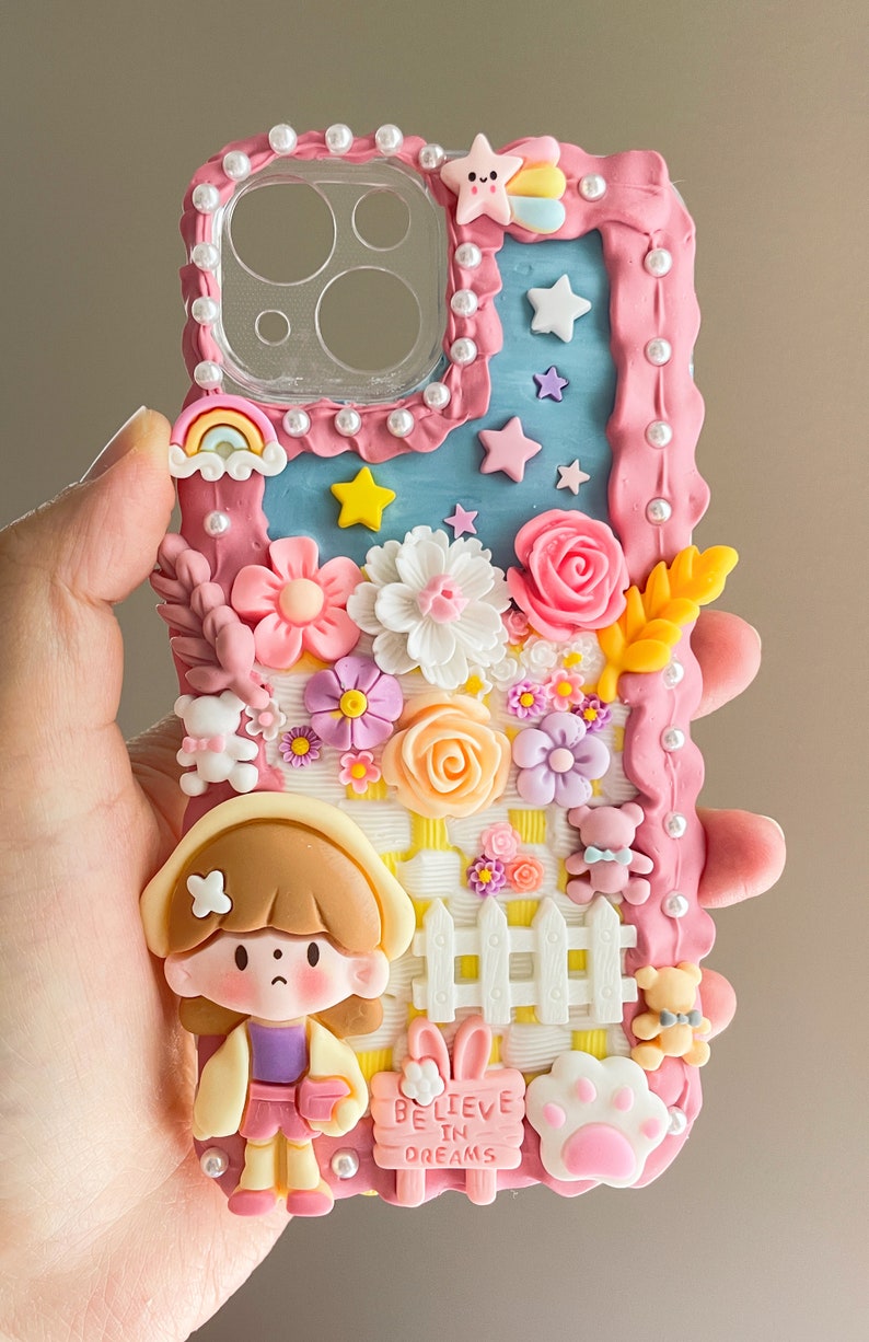 Pu&ograve; includere: Una custodia per telefono rosa e blu decorata con fiori, stelle, un arcobaleno, una recinzione, una ragazza, un coniglio e un'impronta di zampa. La custodia ha il testo "Believe in Dreams" su un cartello giallo.
