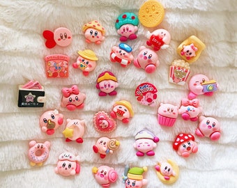 May include: A collection of pink Kirby character-themed charms with various accessories, including hats, bows, and food items. Some charms have Japanese text. The charms are arranged on a white, fluffy surface.