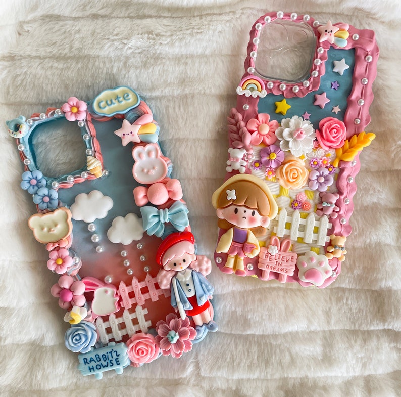 Pu&ograve; includere: Due custodie per telefono rosa e bianche decorate con decorazioni 3D. Una custodia presenta un coniglio dei cartoni animati e il testo "Cute Rabbit House". L'altra custodia presenta una ragazza dei cartoni animati e il testo "Believe in Dreams".