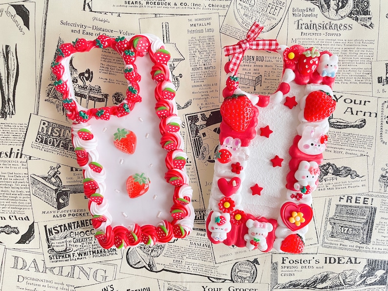 May include: Two white phone cases decorated with red and white frosting, strawberries, and small white bunnies. The cases are shaped like a rectangle with rounded corners.