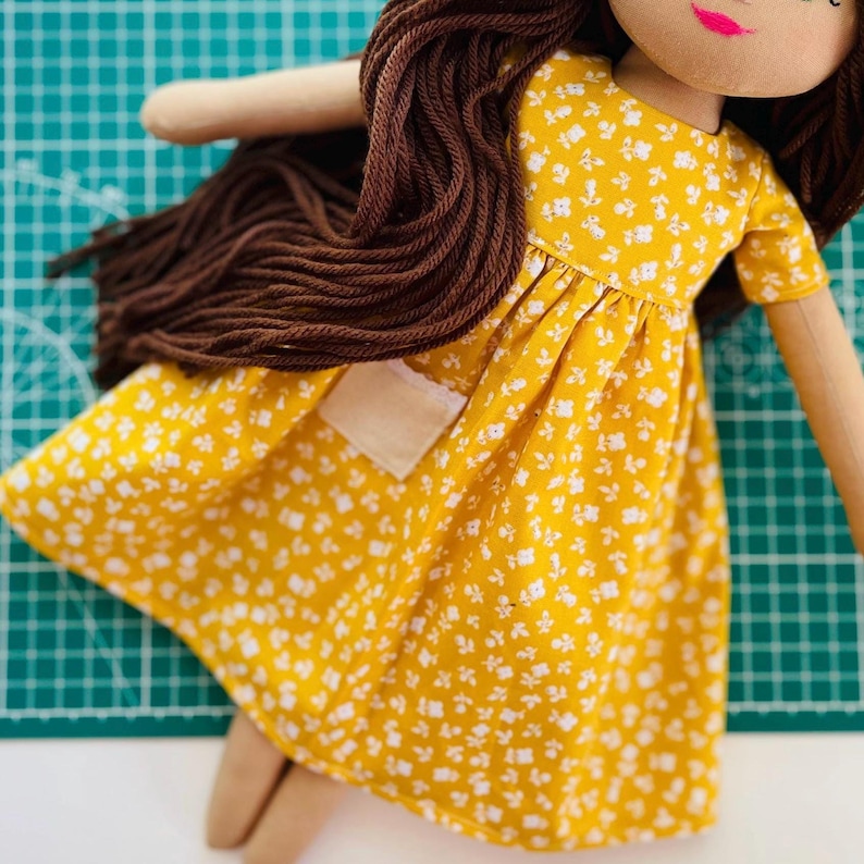 May include: A handmade doll with brown yarn hair and a yellow dress with a white floral pattern. The doll has a light skin tone, pink lips, and a small pocket on the dress. The doll's arms and legs are made of a light beige fabric.