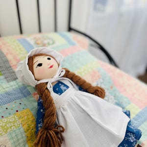 May include: A handmade cloth doll with brown yarn braids, wearing a white bonnet and apron over a blue floral dress. The doll has a painted face with rosy cheeks and is lying on a patchwork quilt.