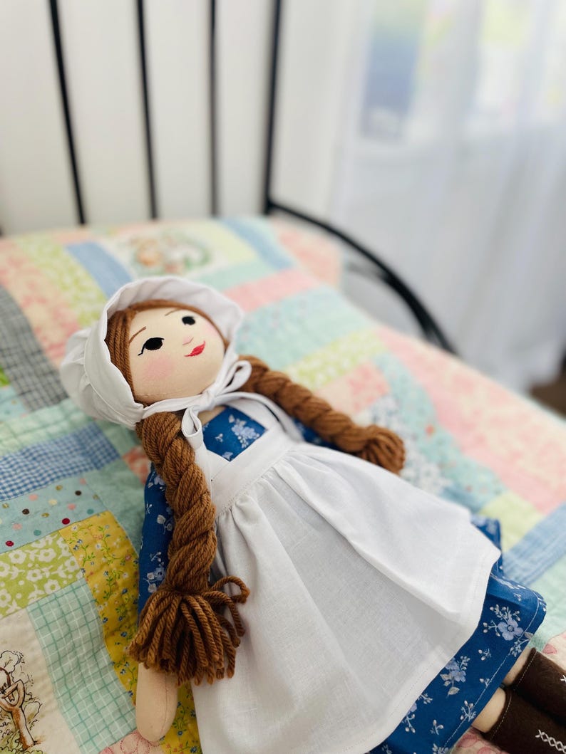 May include: A handmade cloth doll with brown yarn braids, wearing a white bonnet and apron over a blue floral dress. The doll has a painted face with rosy cheeks and is lying on a patchwork quilt.