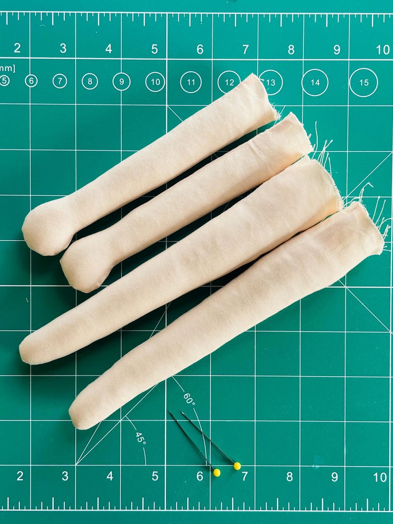 May include: Four beige fabric doll limbs and two sewing pins with yellow heads are arranged on a green cutting mat. The limbs are cylindrical with rounded ends. The pins are positioned near the limbs, suggesting a sewing project.