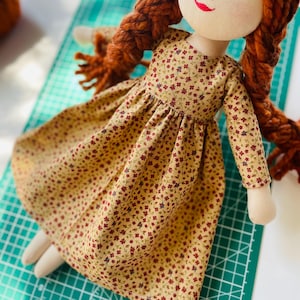 May include: A handmade fabric doll with a cream-colored face, red yarn braids, and a floral print dress. The dress is tan with small red and purple flowers. The doll has a painted face with a red smile and green eyes.