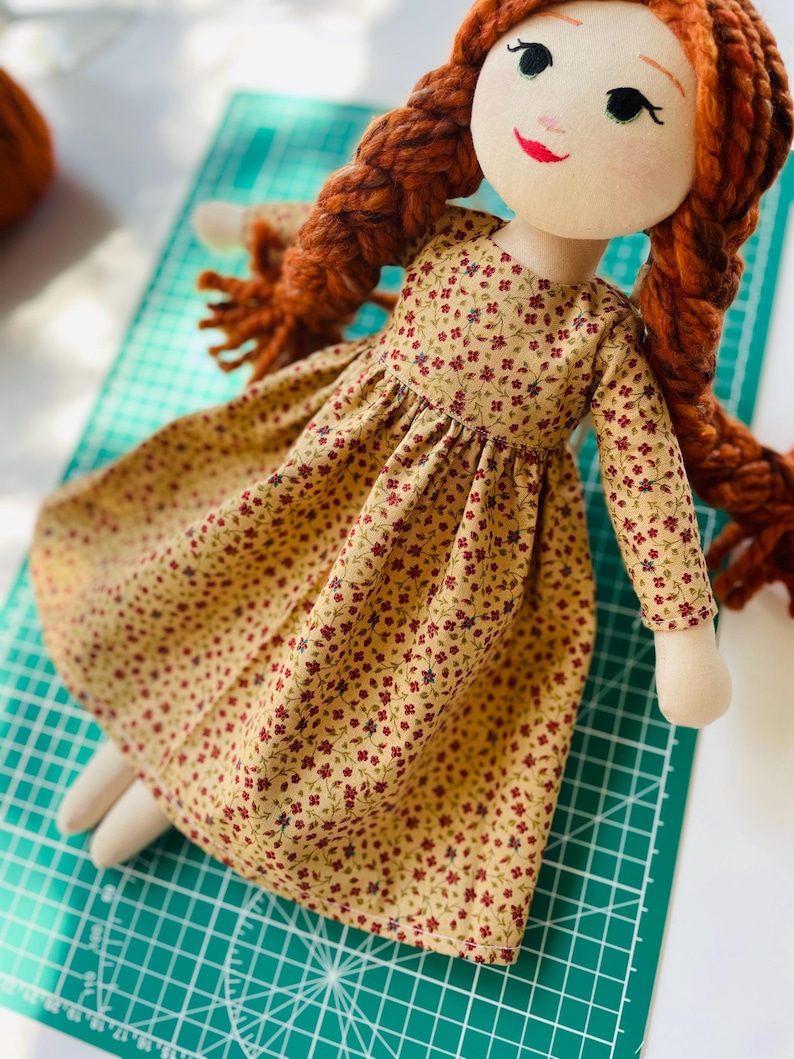 May include: A handmade fabric doll with a cream-colored face, red yarn braids, and a floral print dress. The dress is tan with small red and purple flowers. The doll has a painted face with a red smile and green eyes.