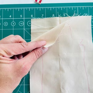May include: A hand holding a piece of light beige fabric with a pink outline of a pattern, possibly for sewing. The fabric is on a green cutting mat with measurements in inches and millimeters.