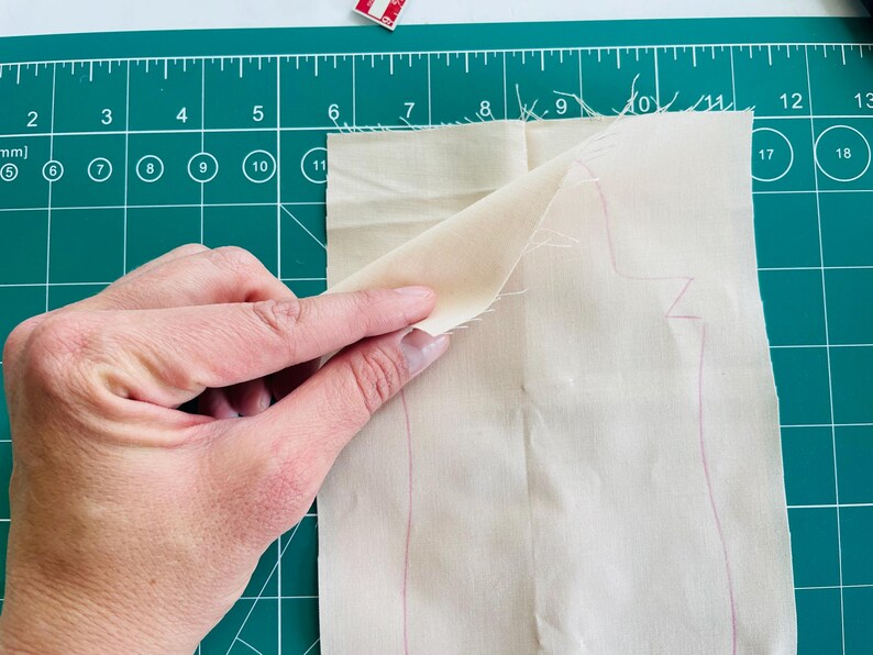 May include: A hand holding a piece of light beige fabric with a pink outline of a pattern, possibly for sewing. The fabric is on a green cutting mat with measurements in inches and millimeters.