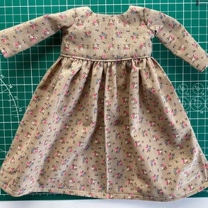 May include: A miniature dress with long sleeves, featuring a beige fabric adorned with a delicate floral pattern in pink and blue. The dress has a gathered skirt and a round neckline, perfect for a doll or small figurine.