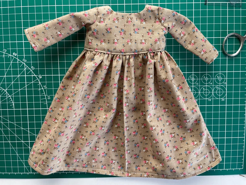 May include: A miniature dress with long sleeves, featuring a beige fabric adorned with a delicate floral pattern in pink and blue. The dress has a gathered skirt and a round neckline, perfect for a doll or small figurine.