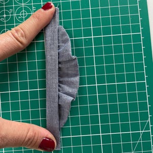Może przedstawiać: Zbliżenie szarego kawałka materiału z marszczoną krawędzią, trzymanego na zielonej macie do cięcia. Materiał jest złożony, tworząc efekt warstwowy. Obraz podkreśla fakturę i szczeg&oacute;ły materiału.