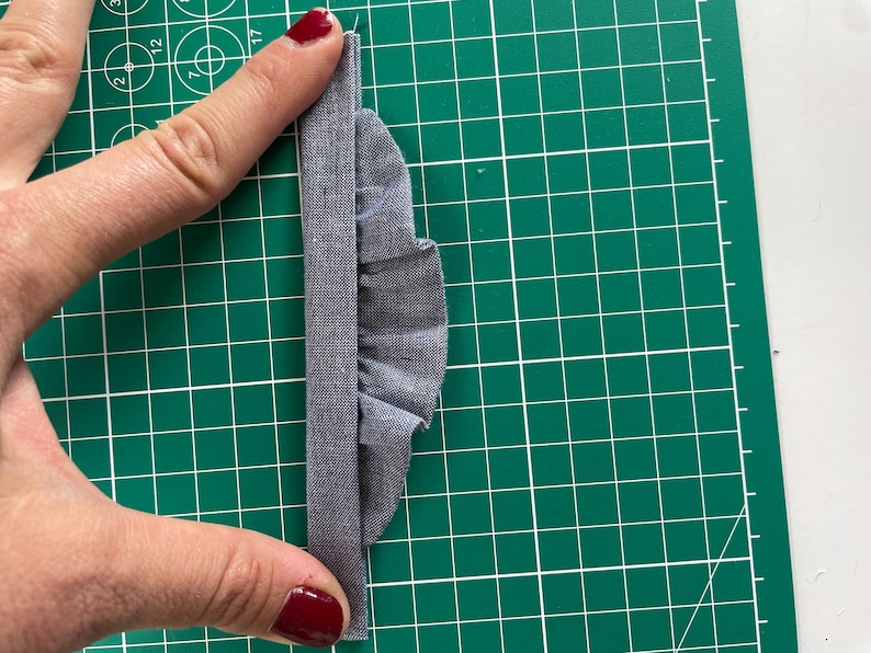 Może przedstawiać: Zbliżenie szarego kawałka materiału z marszczoną krawędzią, trzymanego na zielonej macie do cięcia. Materiał jest złożony, tworząc efekt warstwowy. Obraz podkreśla fakturę i szczeg&oacute;ły materiału.