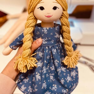 May include: A handmade doll with a fabric face, blonde yarn braids, and a blue floral dress. The doll has embroidered facial features and is held by a person. The doll's dress has a white floral pattern.