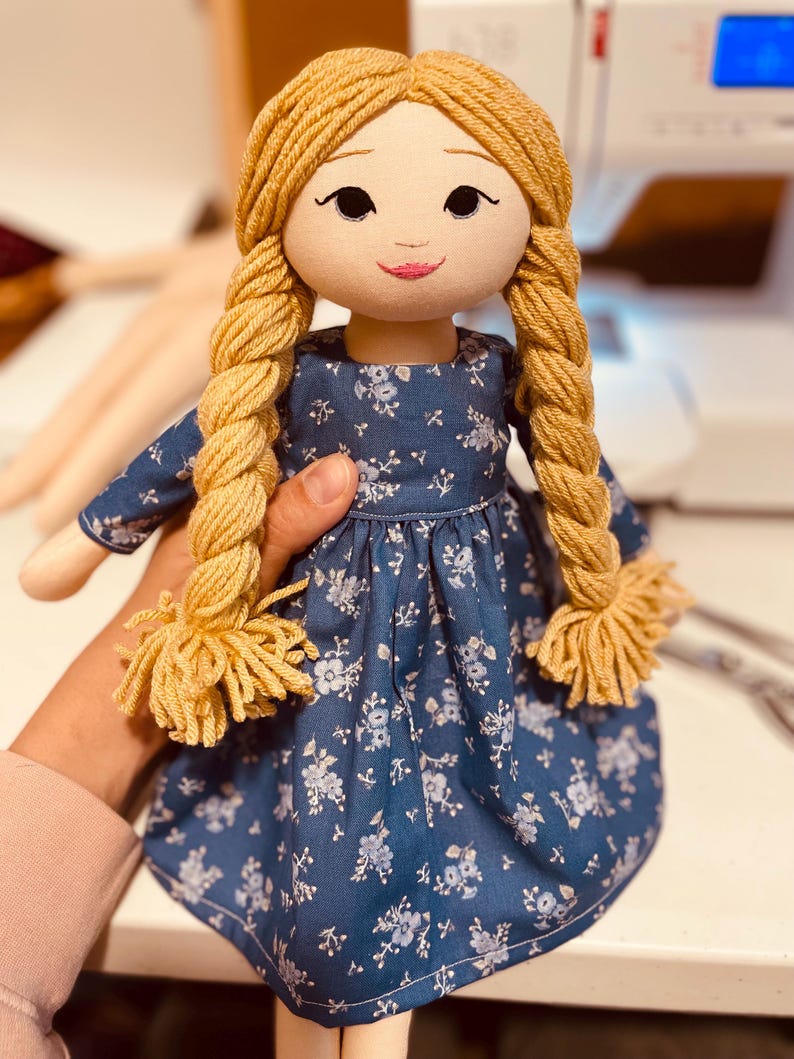 May include: A handmade doll with a fabric face, blonde yarn braids, and a blue floral dress. The doll has embroidered facial features and is held by a person. The doll's dress has a white floral pattern.