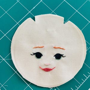 May include: A round, beige fabric face with embroidered features. Black eyes with long lashes, a small pink nose, and a red smiling mouth are stitched onto the fabric. The eyebrows are orange. The fabric has a unique shape with a notched top.
