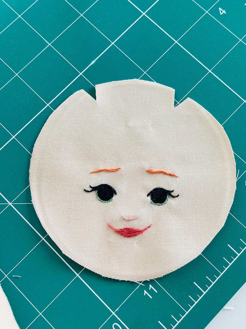 May include: A round, beige fabric face with embroidered features. Black eyes with long lashes, a small pink nose, and a red smiling mouth are stitched onto the fabric. The eyebrows are orange. The fabric has a unique shape with a notched top.