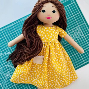 May include: A handmade doll with brown yarn hair and a yellow floral dress. The doll has a light skin tone, a pink mouth, and black eyes. The dress has a small pocket and short sleeves. The doll's arms and legs are made of a beige fabric.