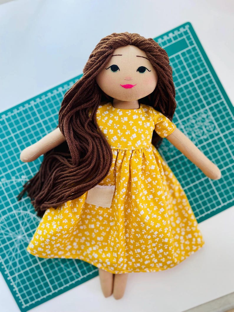 May include: A handmade doll with brown yarn hair and a yellow floral dress. The doll has a light skin tone, a pink mouth, and black eyes. The dress has a small pocket and short sleeves. The doll's arms and legs are made of a beige fabric.
