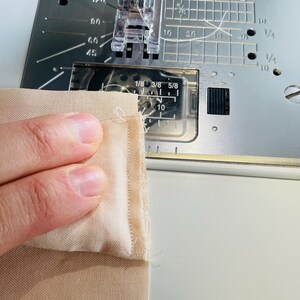 May include: Close-up of a sewing machine in operation, stitching together two pieces of beige fabric. The machine's needle is piercing the fabric, creating a seam. The machine has measurement markings in inches.