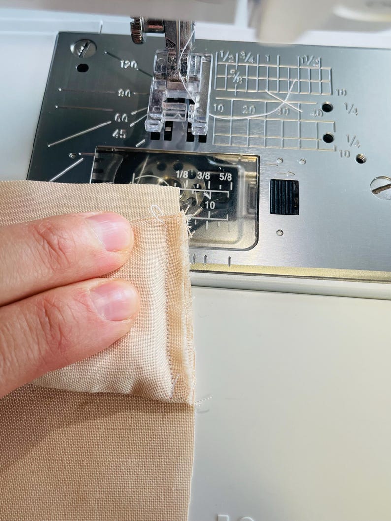 May include: Close-up of a sewing machine in operation, stitching together two pieces of beige fabric. The machine's needle is piercing the fabric, creating a seam. The machine has measurement markings in inches.