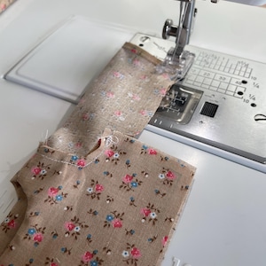 May include: A sewing machine stitching floral fabric. The fabric is tan with a pattern of pink, blue, and white flowers. The machine is white with silver components. The fabric is being sewn in a straight line.